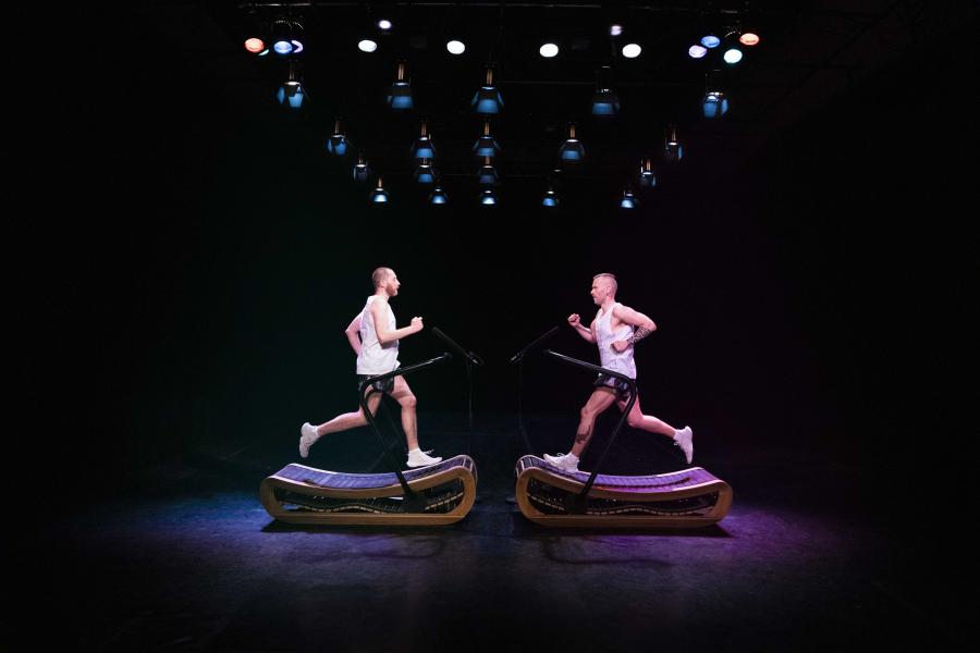 You can see two treadmills opposite each other on which one person is running, the two people are running towards each other. The background is black, the two persons are illuminated in purple and blue